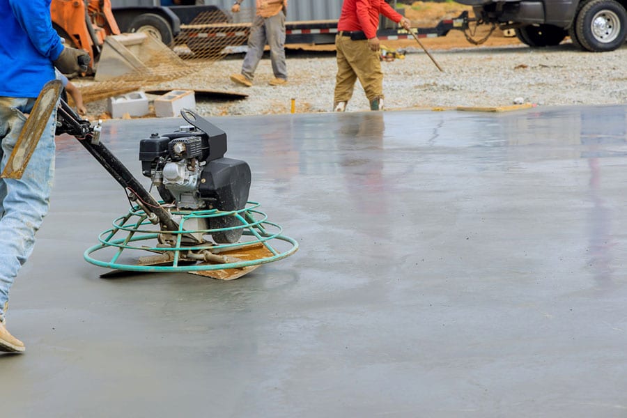 A worker from Metro Detroit Concrete Contractors uses a power trowel to smooth wet concrete on a construction site, while others work in the background near equipment and construction materials.