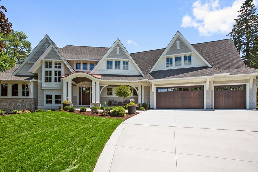A large, modern suburban house with gabled roofs, gray siding, stone accents, and a three-car garage with wooden doors. The neatly manicured lawn features a curved concrete driveway in front, perfect for Metro Detroit living.