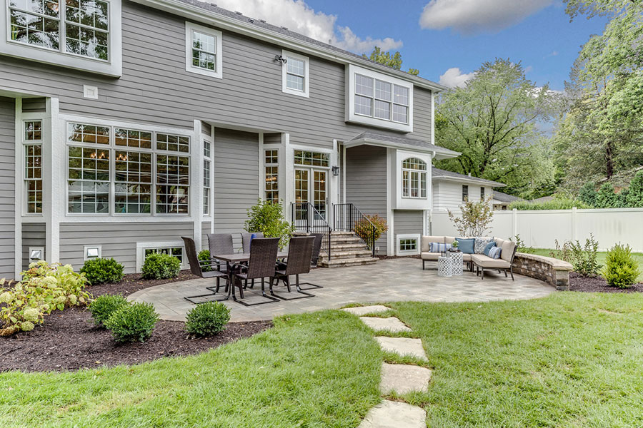 A modern gray house with large windows overlooks a backyard patio featuring outdoor dining and seating areas, surrounded by landscaped gardens, green lawn, and a stone path—perfect inspiration for concrete patios in Metro Detroit.