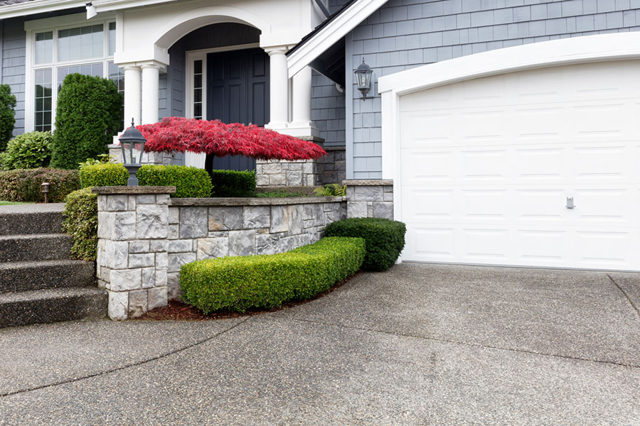 Front view of a Metro Detroit house with a gray stone retaining wall, green shrubs, a red-leafed bush, black front door, white garage door, and decorative concrete driveway and pathway.