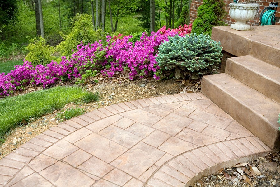 Curved decorative concrete patio with adjacent steps, bordered by vibrant pink flowers, small evergreen shrubs, and green grass, next to a brick wall and a white planter. Trees and greenery are visible in the background of this Metro Detroit home.