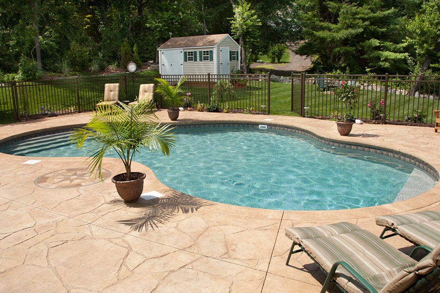 Sunny backyard with an in-ground swimming pool, surrounded by stamped concrete patio and lounge chairs. Perfect for enjoying backyard pools in Metro Detroit, with green grass, trees, potted plants, and a small shed in the background.