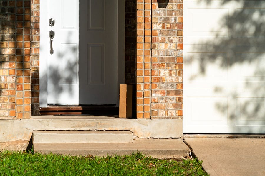 A cardboard package sits on the porch in front of a white door, next to a garage, at a brick house on a sunny day. Subtle signs of porch foundation issues can be seen where the front porch is pulling away from the main structure.