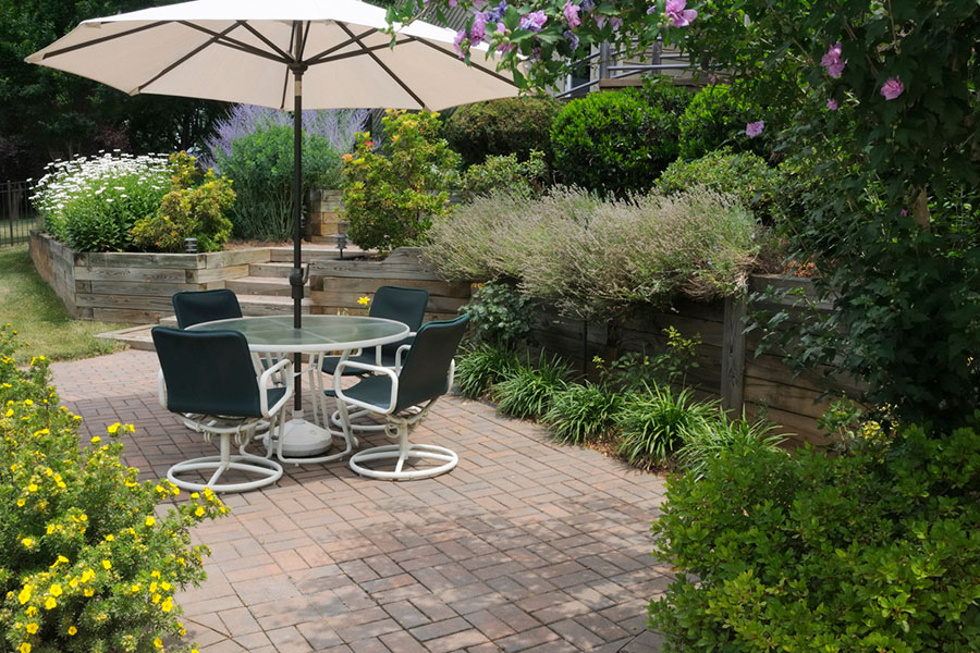 A patio with a round glass table, four chairs, and a large umbrella is set atop beautiful spring brick paving, surrounded by lush greenery, blooming flowers, and raised garden beds.