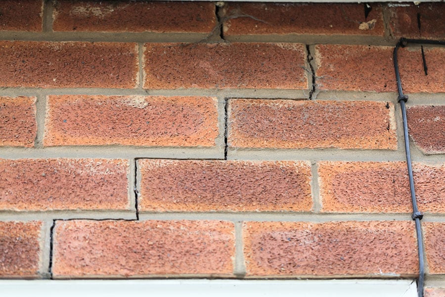 Structural cracks running through the mortar and bricks of an exterior wall, extending from the window and doorway.